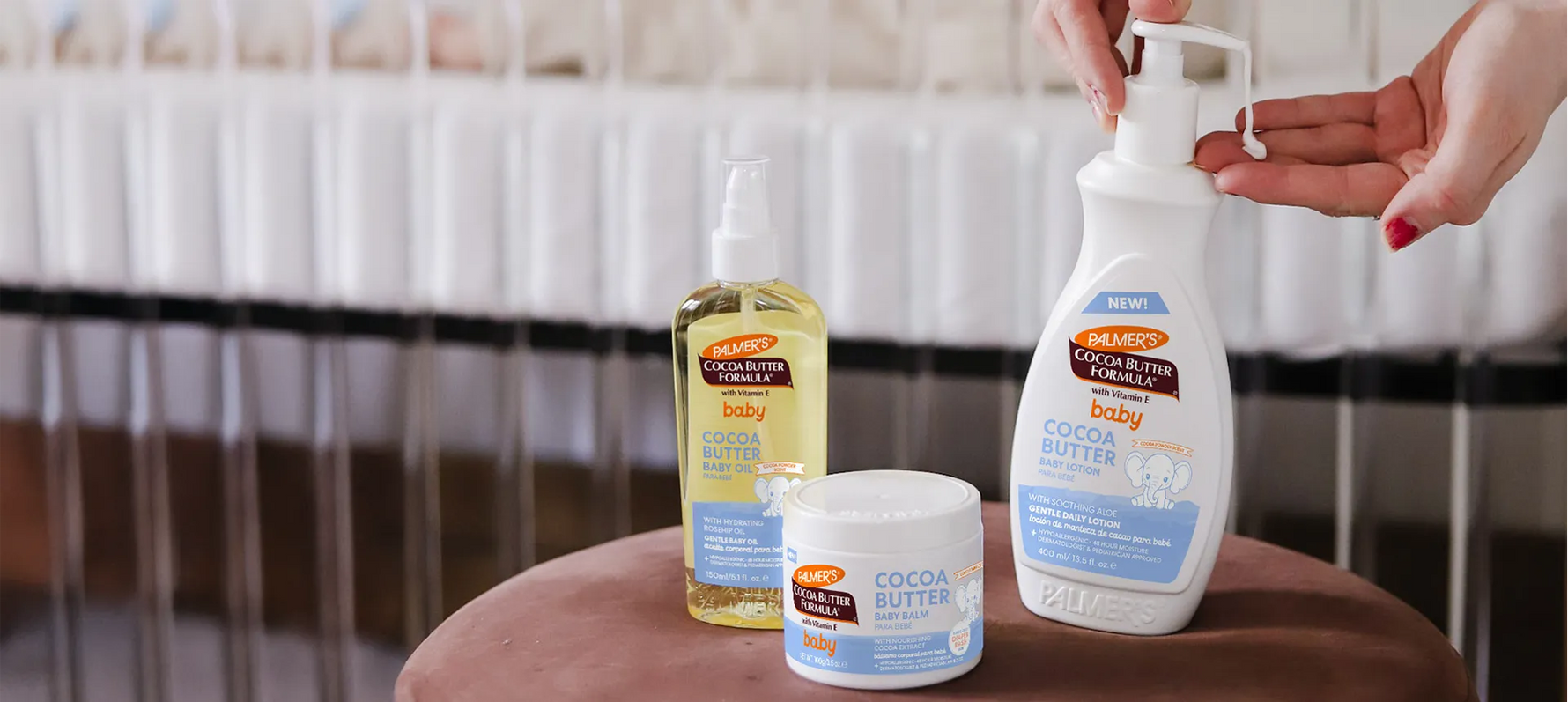 Palmer's Cocoa Butter Baby Skin Care products on ottoman in front of crib with lotion being pumped out into woman's hand