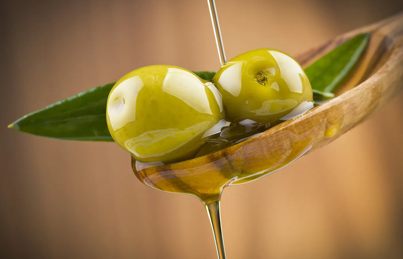 Two green olives on a wooden spoon with olive oil dripping, against a blurred background.