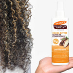 Palmer's Cocoa Butter Formula leave-in conditioner bottle held by a hand next to curly hair.