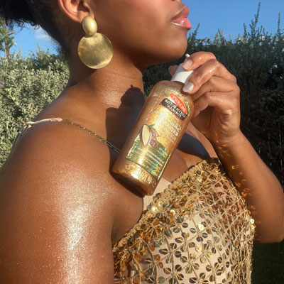 Black woman in gold dress holding Palmer's Shimmer Radiance Body Oil, her favorite holiday skin care product