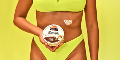 Woman in yellow bikini holding Palmer's Brazilian Coco Cream for minimizing cellulite in front of her stomach with heart made of lotion on her abdomen