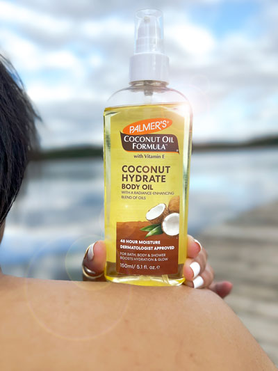 Woman holding Palmer's Coconut Hydrate Body Oil, part of a hydrating skin care routine, on her bare shoulder outdoors 